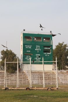 A cricket scoreboard in Hyderabad, Pakistan, with birds flying above under a clear sky.