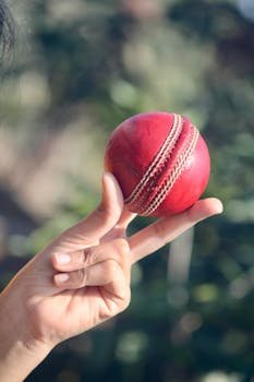 Detailed close-up of a hand skillfully holding a red leather cricket ball outdoors.