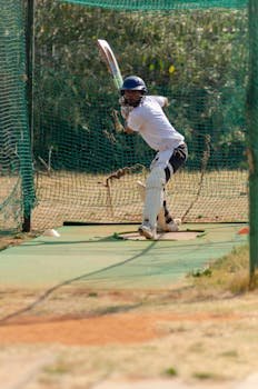 A cricket player practicing a sweep shot in outdoor nets under bright daylight.