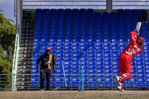 A cricket player in red batting during a match in an empty stadium with blue seats.