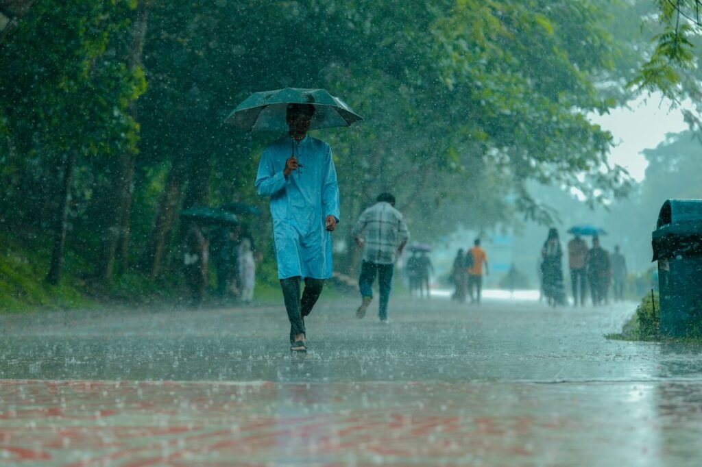 A man in blue walking under heavy rain with umbrella on a tree-lined path.