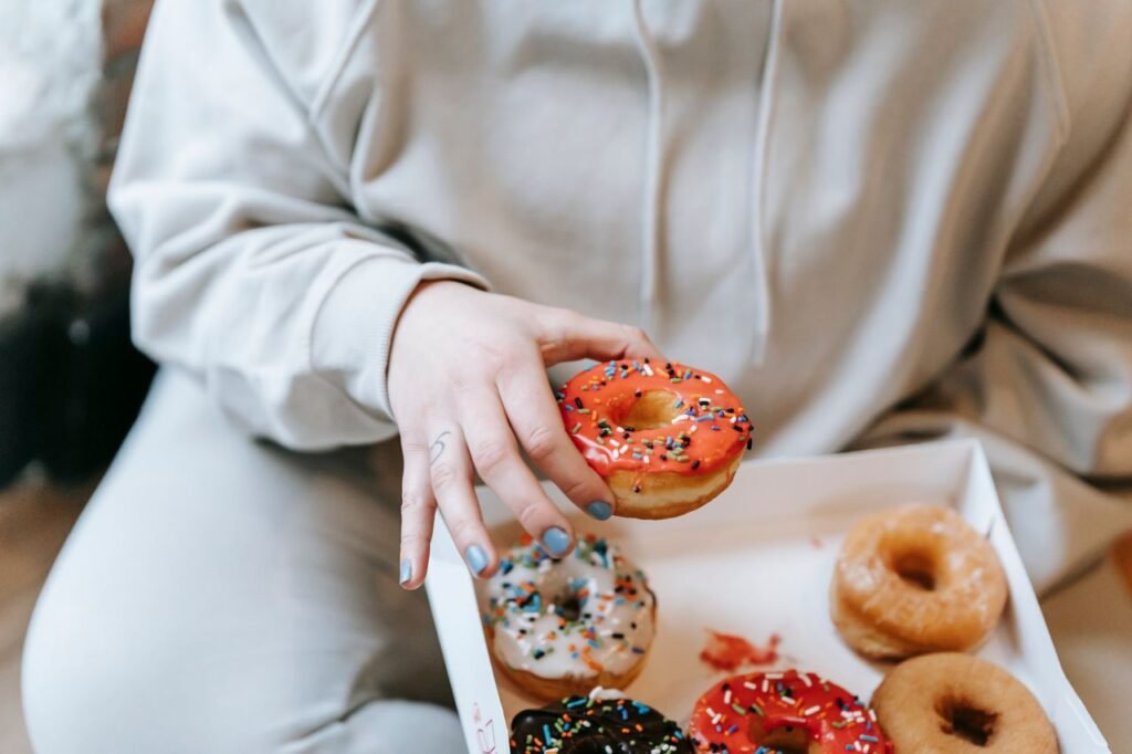 Crop unrecognizable plump female in comfy clothes picking yummy sweet donut from carton box while sitting on floor in light room