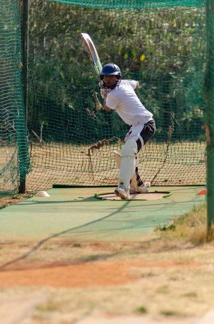 A cricket player practicing a sweep shot in outdoor nets under bright daylight.