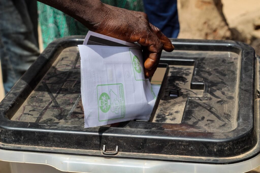 A close-up shot of a hand placing a paper ballot into a transparent voting box outdoors.