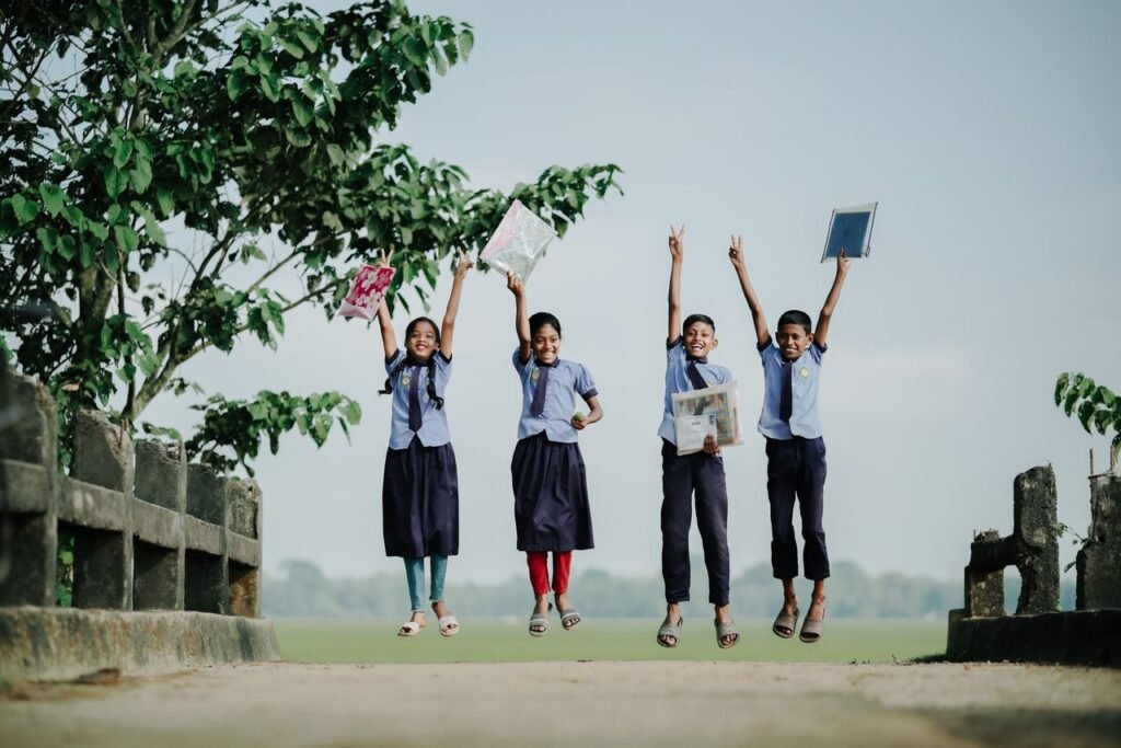 Four schoolchildren in uniforms joyfully jump on a bridge, holding books and papers, with trees in the background.